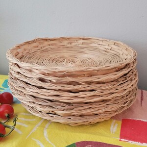 May include: A stack of five round, shallow woven baskets made from light tan natural reeds. Each basket features a circular base with a spiraling weave pattern and a thin, flexible rim. The baskets are stacked vertically, creating a uniform profile.