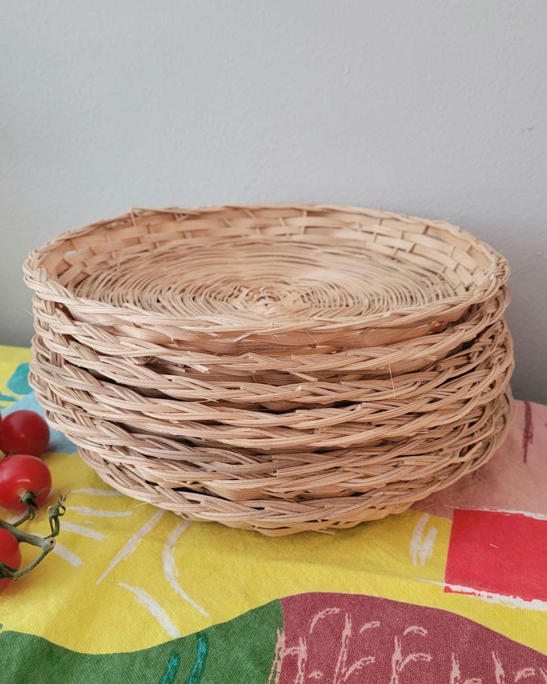 May include: A stack of five round, shallow woven baskets made from light tan natural reeds. Each basket features a circular base with a spiraling weave pattern and a thin, flexible rim. The baskets are stacked vertically, creating a uniform profile.