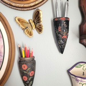 May include: A collection of decorative wall-mounted holders and art pieces. Includes a gold butterfly, two black holders with floral and landscape designs, and a vase with colored pencils and paintbrushes.