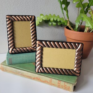 May include: Two rectangular picture frames with a metallic copper-colored rope-style border and a black inner edge. One frame is oriented vertically and the other horizontally, both featuring a plain tan insert within the frame opening.