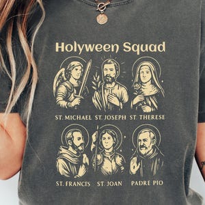 May include: Dark gray t-shirt with the text "Holyween Squad" and illustrations of six saints. The shirt also features the text "ST. MICHAEL ST. JOSEPH ST. THERESE" and "ST. FRANCIS ST. JOAN PADRE PIO". The shirt is from Comfort Colors.