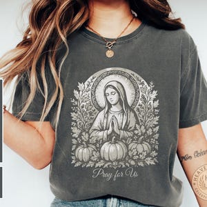 May include: Dark grey t-shirt featuring a religious illustration of a woman with a halo, praying hands, and pumpkins. The text "Pray for Us" is below the image. The shirt has the words "Comfort Colors 1717 Pepper" printed on the side.