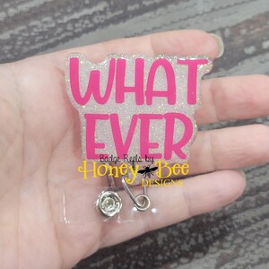 May include: A white glitter badge reel with a pink "WHAT EVER" text overlay. The badge reel has a silver clip and a clear plastic backing. The text "Badge Reels by Honey Bee Designs" is printed below the text.