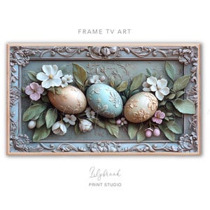 May include: A framed artwork featuring three pastel-colored eggs nestled in a bed of green leaves and delicate white flowers. The eggs are decorated with intricate patterns and the frame is ornate with a gold finish.