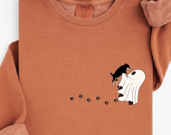Puede incluir: Una sudadera de color óxido con cuello redondo y mangas largas. El diseño presenta un perro negro saltando sobre un fantasma blanco, con huellas de patas. La palabra "YAM" está impresa en la parte inferior derecha.