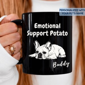 Könnte beinhalten: Ein schwarzer Keramikbecher mit einer weißen Strichzeichnung einer liegenden Französischen Bulldogge. Der Text lautet Emotional Support Potato und Buddy. Ein blaues Etikett oben rechts zeigt PERSONALIZED WITH YOUR PET'S NAME.