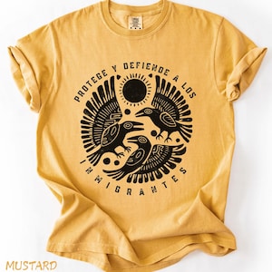 May include: Mustard-colored t-shirt with a black graphic featuring three stylized birds, a sun, and the text "PROTEGE Y DEFIENDE A LOS INMIGRANTES." The shirt has short sleeves and a crew neck. The word "MUSTARD" is printed at the bottom.