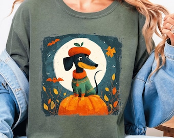 Op de afbeelding: Mosgroen sweatshirt met een Halloween-thema. De afbeelding toont een teckel met een baret en trui, zittend op een oranje pompoen voor een volle maan. Vleermuizen en herfstbladeren zijn ook afgebeeld.