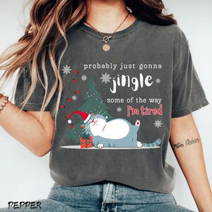 Puede incluir: Camiseta gris oscuro con un gr&aacute;fico de gato navide&ntilde;o. El gato lleva un gorro de Pap&aacute; Noel y est&aacute; acostado junto a un &aacute;rbol de Navidad. El texto de la camiseta dice "probably just gonna jingle some of the way I'm tired."