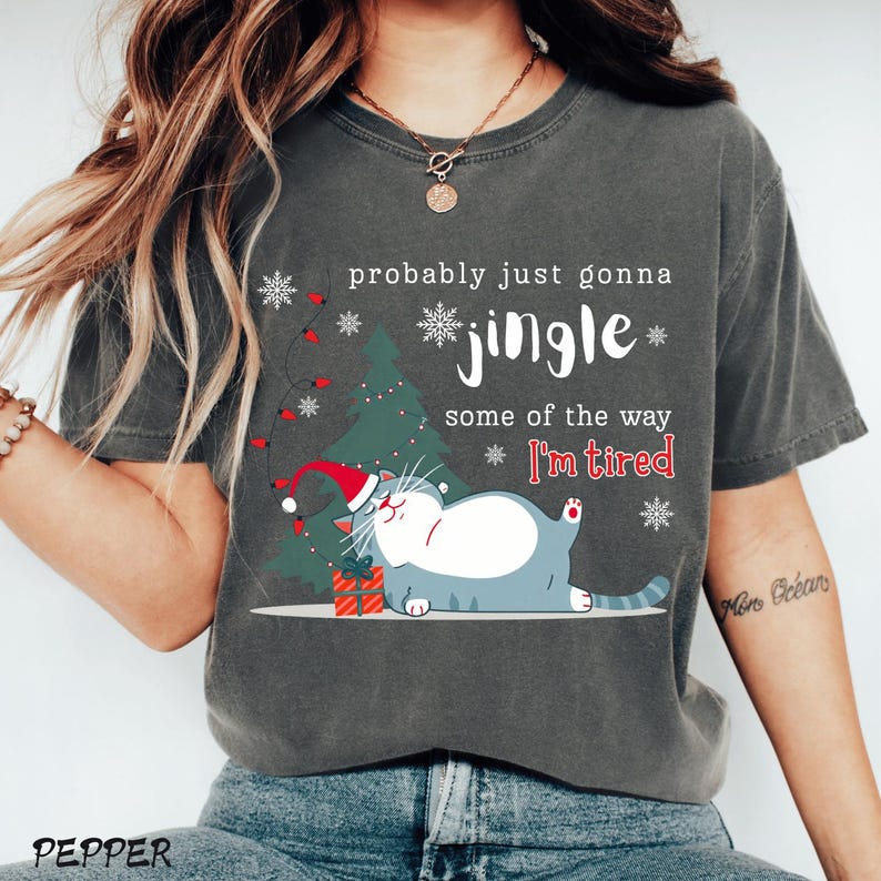 Puede incluir: Camiseta gris oscuro con un gr&aacute;fico de gato navide&ntilde;o. El gato lleva un gorro de Pap&aacute; Noel y est&aacute; acostado junto a un &aacute;rbol de Navidad. El texto de la camiseta dice "probably just gonna jingle some of the way I'm tired."