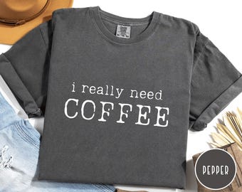 May include: A dark gray t-shirt with the text "i really need COFFEE" in white, centered on the front. The shirt has short, rolled-up sleeves and a crew neck. The shirt is displayed on a white surface.