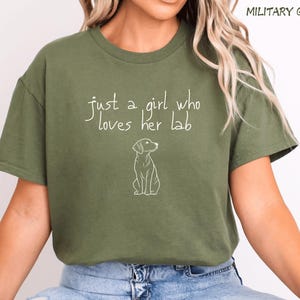 Puede incluir: Camiseta verde militar con el texto "just a girl who loves her lab" y un contorno blanco de un perro. La camiseta es de color liso y tiene mangas cortas.