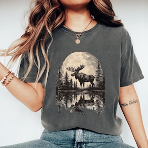 May include: A charcoal grey t-shirt with a graphic print of a moose standing in front of a full moon reflected in a lake. The moose is silhouetted against the moon and the trees behind it. The text "More Oceans" is written on the right sleeve of the shirt.