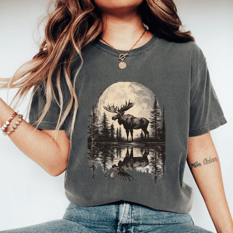 May include: A charcoal grey t-shirt with a graphic print of a moose standing in front of a full moon reflected in a lake. The moose is silhouetted against the moon and the trees behind it. The text "More Oceans" is written on the right sleeve of the shirt.