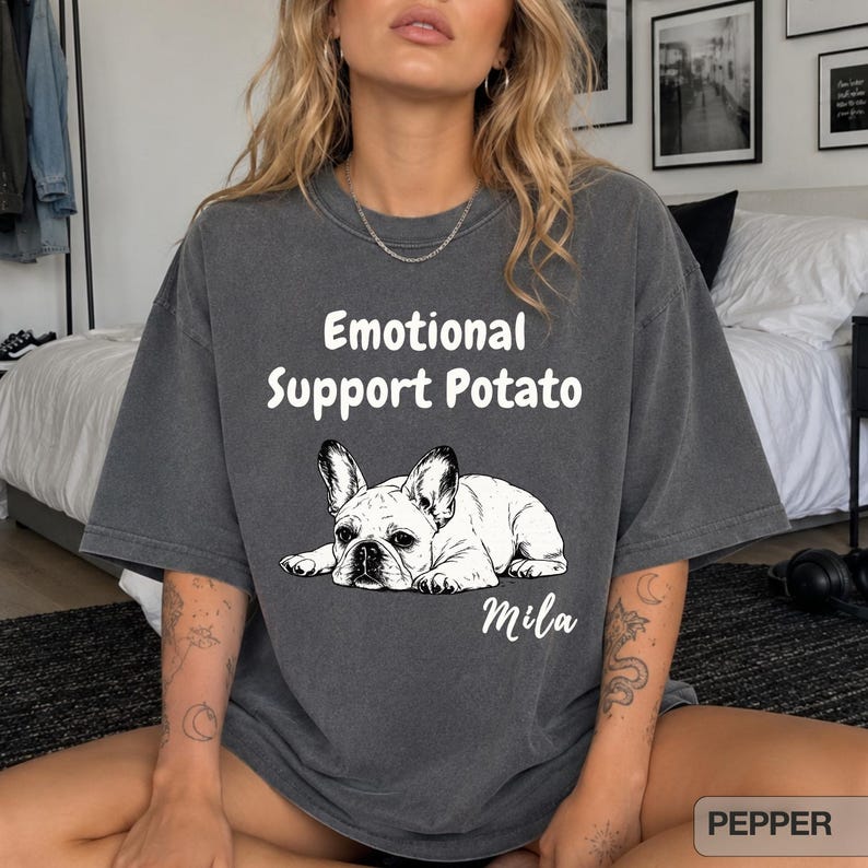 May include: A charcoal gray short-sleeve t-shirt featuring a white graphic of a French Bulldog lying down. Above the dog, the text reads "Emotional Support Potato" in a white sans-serif font, with the name "Mila" printed in white script below.