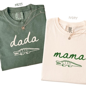 Op de afbeelding: Twee t-shirts, één mosgroen en één ivoor, met de woorden "dada" en "mama" in cursief schrift. Elk shirt heeft een krokodilafbeelding. Beide shirts hebben een zilveren ketting.