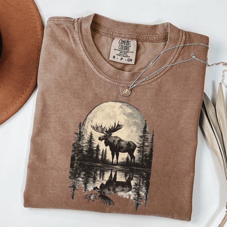 May include: A brown t-shirt with a graphic of a moose standing in front of a full moon and a reflection of the moose in a lake. The text "COMFORT COLORS" is on the tag.