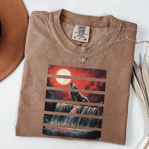 May include: A brown t-shirt with a graphic print of a wolf howling at the moon in front of a waterfall. The graphic is in a layered style with red, orange, and black colors. The t-shirt has a tag that says "Comfort Colors" and "S - P - CH".