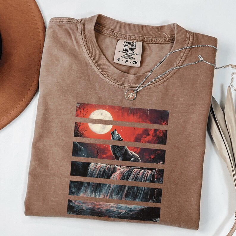 May include: A brown t-shirt with a graphic print of a wolf howling at the moon in front of a waterfall. The graphic is in a layered style with red, orange, and black colors. The t-shirt has a tag that says "Comfort Colors" and "S - P - CH".