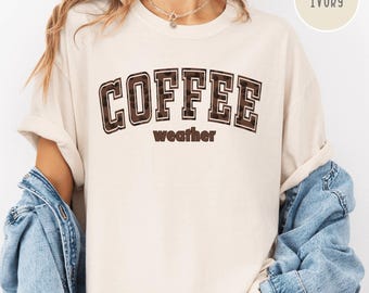 Könnte beinhalten: Elfenbeinfarbenes T-Shirt mit dem Schriftzug "COFFEE weather" in braunen Blockbuchstaben. Das Shirt hat kurze Ärmel und wird mit einer Jeansjacke kombiniert. Die Farbe "IVORY" ist in einem Kreis oben rechts angegeben.