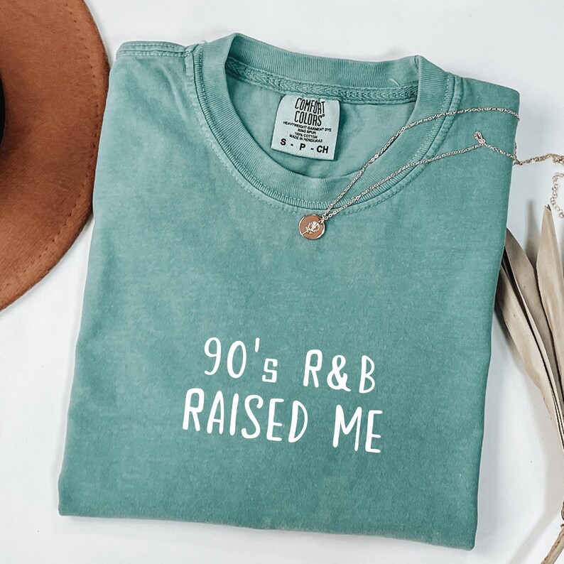 Puede incluir: Una camiseta azul turquesa con el texto "90's R&B RAISED ME" en letras blancas.