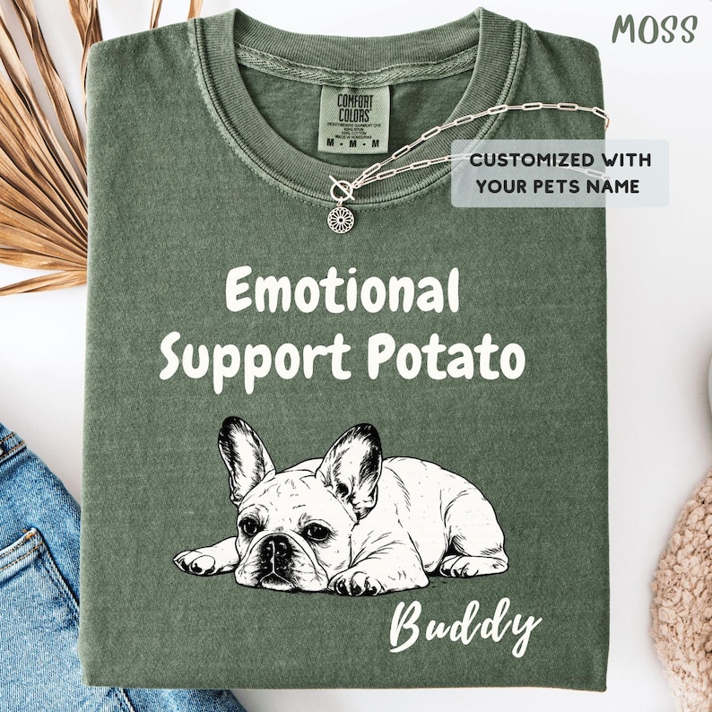 May include: A moss green t-shirt featuring a white line-art illustration of a French Bulldog lying down. The text reads "Emotional Support Potato" above the dog and "Buddy" below. A label states "CUSTOMIZED WITH YOUR PETS NAME" and a silver necklace is visible.