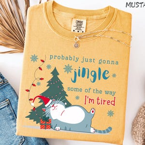 Puede incluir: Camiseta de color mostaza con un gr&aacute;fico de un gato con gorro de Pap&aacute; Noel, un &aacute;rbol de Navidad y el texto "probably just gonna jingle some of the way I'm tired." Un collar plateado est&aacute; sobre la camiseta.