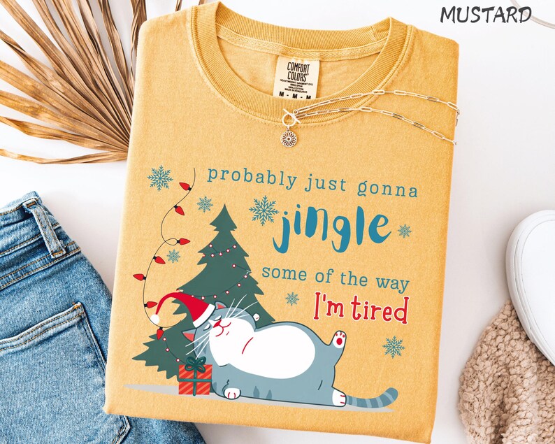 Puede incluir: Camiseta de color mostaza con un gr&aacute;fico de un gato con gorro de Pap&aacute; Noel, un &aacute;rbol de Navidad y el texto "probably just gonna jingle some of the way I'm tired." Un collar plateado est&aacute; sobre la camiseta.