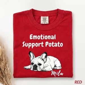 May include: A red crew neck t-shirt featuring a black and white line art illustration of a French Bulldog lying down. The text "Emotional Support Potato" is printed in white above the dog, and the name "Mila" is printed in white below the dog.