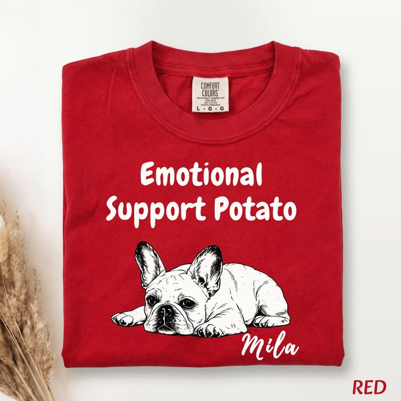 May include: A red crew neck t-shirt featuring a black and white line art illustration of a French Bulldog lying down. The text "Emotional Support Potato" is printed in white above the dog, and the name "Mila" is printed in white below the dog.