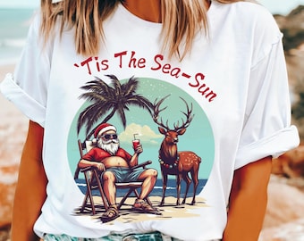 May include: White t-shirt featuring a beach scene with Santa Claus, a reindeer, and a palm tree. Santa is relaxing in a chair and holding a drink. The text "'Tis The Sea-Sun" is printed above the graphic.