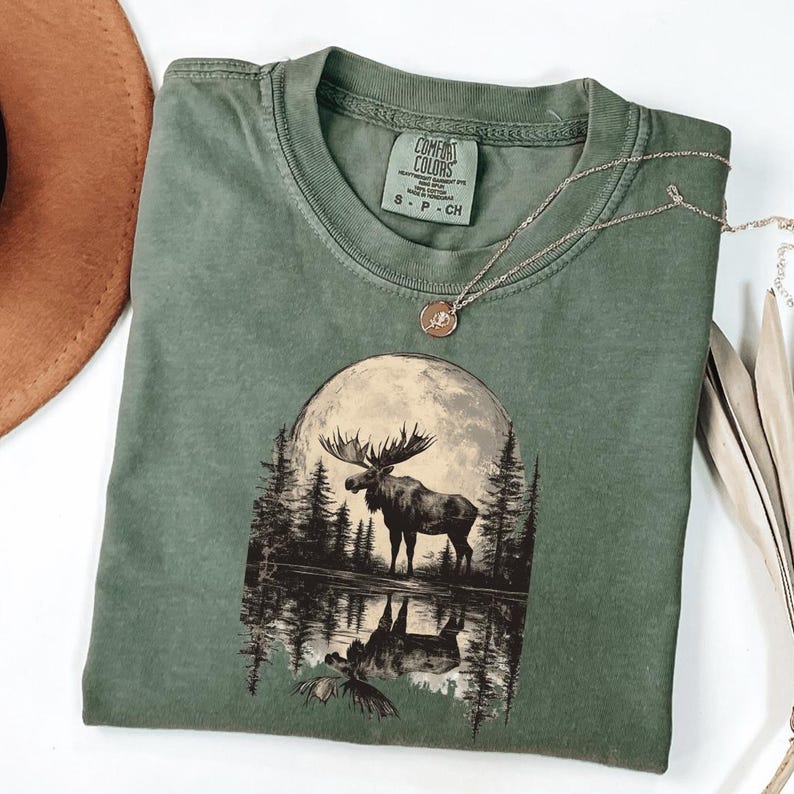 May include: A green t-shirt with a graphic of a moose standing in front of a full moon reflected in a lake. The t-shirt has a tag that says "Comfort Colors" and "S - P - CH".