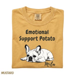 May include: A mustard yellow t-shirt featuring a black line art illustration of a French Bulldog lying down. Above the dog, the text reads Emotional Support Potato in a dark font. The name Mila is printed in a script font below the dog.