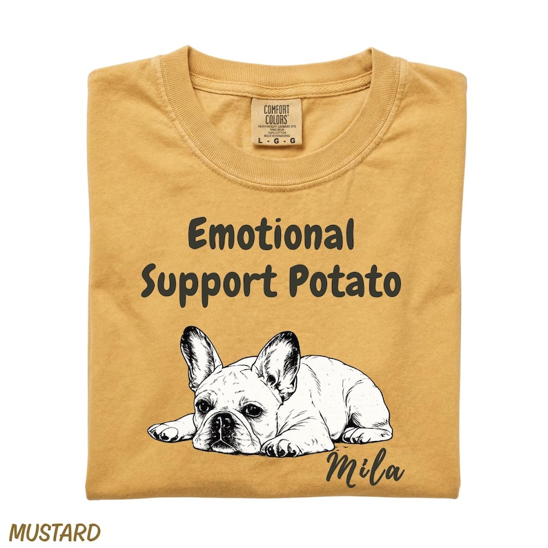 May include: A mustard yellow t-shirt featuring a black line art illustration of a French Bulldog lying down. Above the dog, the text reads Emotional Support Potato in a dark font. The name Mila is printed in a script font below the dog.