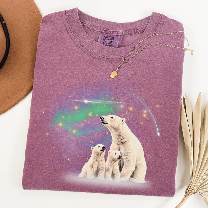 May include: A purple t-shirt with a graphic of three polar bears looking up at a starry sky with a green aurora borealis. The shirt has the text "Comfort Colors" on the tag.