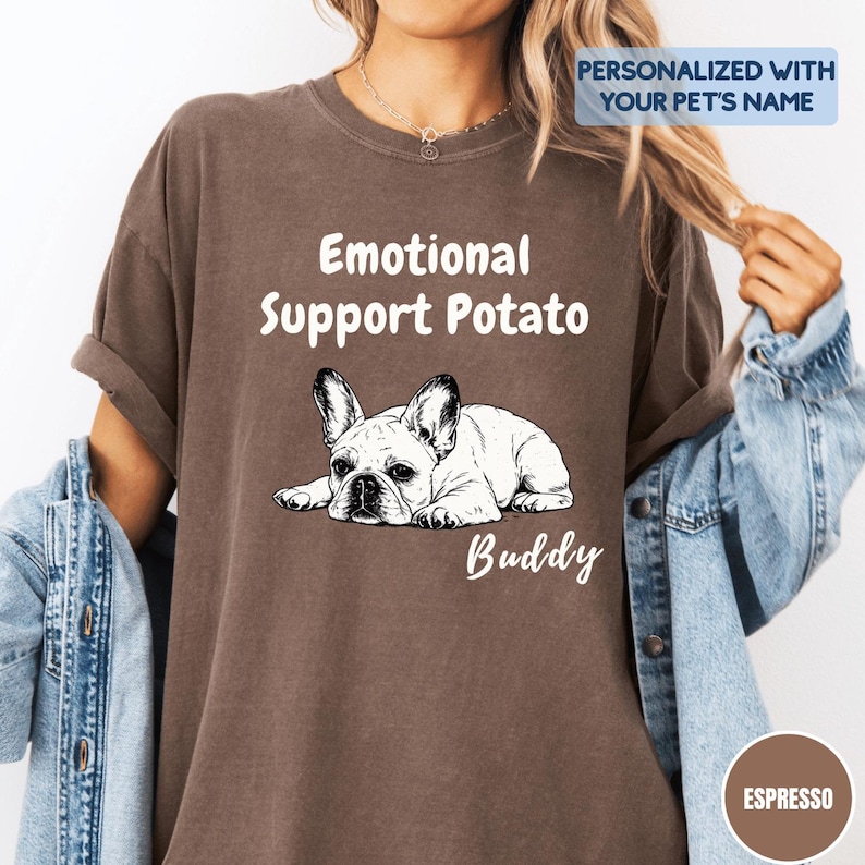 May include: An espresso-colored short-sleeve t-shirt featuring a white graphic of a French Bulldog lying down. The text reads "Emotional Support Potato" above the dog and "Buddy" below. A blue box in the corner says "PERSONALIZED WITH YOUR PET'S NAME."