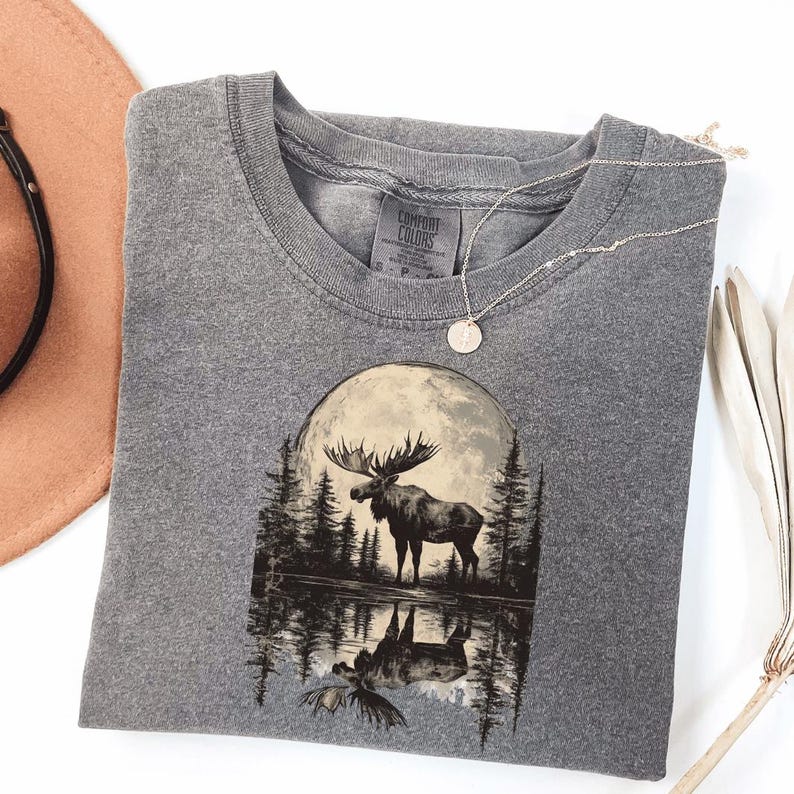 May include: A gray t-shirt with a graphic of a moose standing in a forest with a full moon behind it. The moose is reflected in the water below. The t-shirt has the text "Comfort Colors" on the tag.