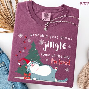 Puede incluir: Camiseta color bayas con un gr&aacute;fico de un gato con gorro de Pap&aacute; Noel, un &aacute;rbol de Navidad y el texto "probably just gonna jingle some of the way I'm tired." Un collar plateado est&aacute; sobre la camiseta.