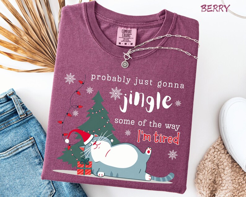 Puede incluir: Camiseta color bayas con un gr&aacute;fico de un gato con gorro de Pap&aacute; Noel, un &aacute;rbol de Navidad y el texto "probably just gonna jingle some of the way I'm tired." Un collar plateado est&aacute; sobre la camiseta.