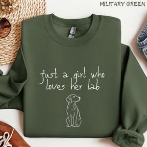 Puede incluir: Sudadera verde militar con el texto "just a girl who loves her lab" y un dibujo lineal blanco de un Labrador Retriever. La sudadera es de color liso y tiene cuello redondo.