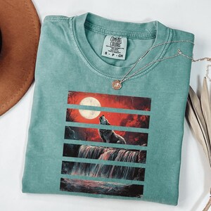 May include: Teal blue t-shirt with a graphic print of a wolf howling at the moon in front of a waterfall. The graphic is in a vintage style with red, orange, and black colors. The t-shirt has the text "COMFORT COLORS" and "S - P - CH" printed on the tag.