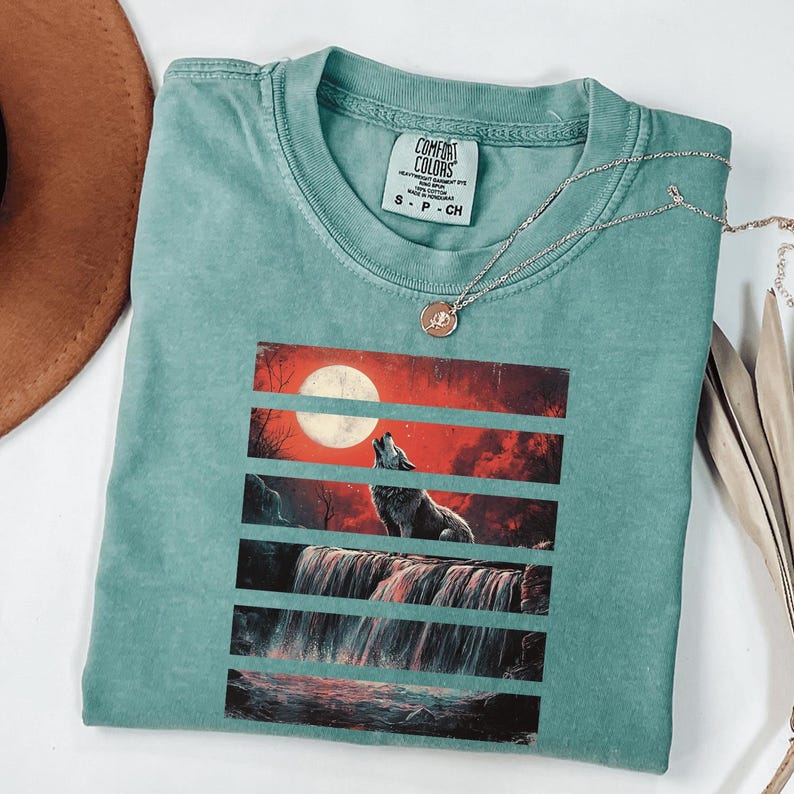 May include: Teal blue t-shirt with a graphic print of a wolf howling at the moon in front of a waterfall. The graphic is in a vintage style with red, orange, and black colors. The t-shirt has the text "COMFORT COLORS" and "S - P - CH" printed on the tag.