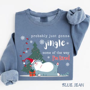 Puede incluir: Sudadera azul vaquera con un gr&aacute;fico de gato navide&ntilde;o. El gato lleva un gorro de Pap&aacute; Noel y est&aacute; junto a un &aacute;rbol de Navidad y un regalo. El texto dice "probably just gonna jingle some of the way I'm tired."
