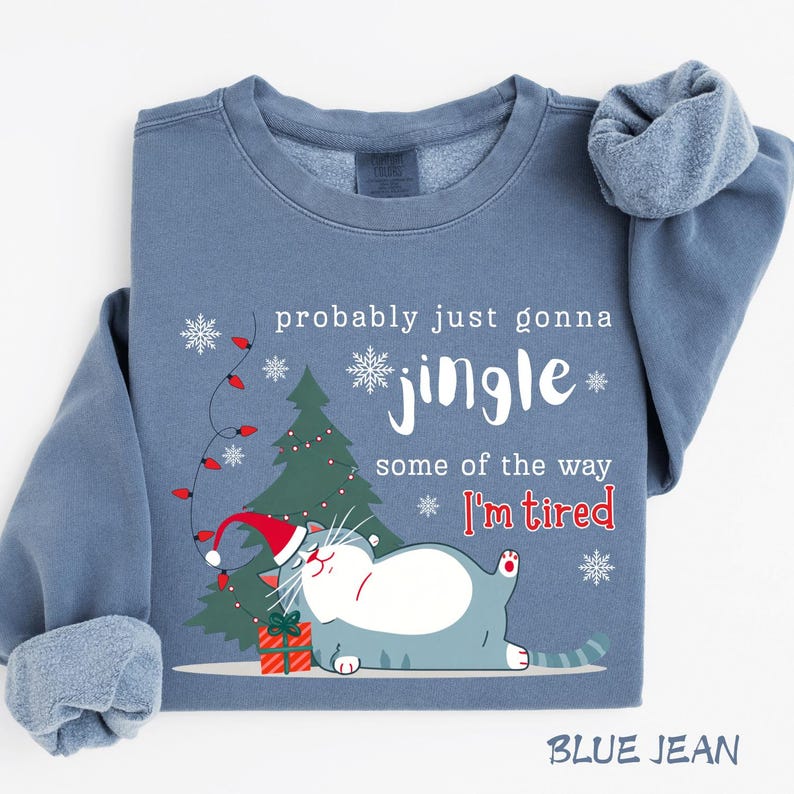 Puede incluir: Sudadera azul vaquera con un gr&aacute;fico de gato navide&ntilde;o. El gato lleva un gorro de Pap&aacute; Noel y est&aacute; junto a un &aacute;rbol de Navidad y un regalo. El texto dice "probably just gonna jingle some of the way I'm tired."
