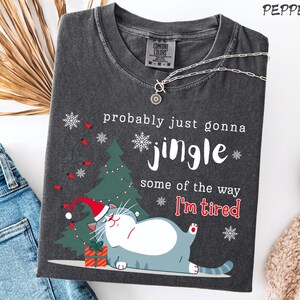 Puede incluir: Camiseta gris oscuro con un gr&aacute;fico de gato navide&ntilde;o. El gato lleva un gorro de Pap&aacute; Noel y est&aacute; junto a un &aacute;rbol de Navidad y un regalo. El texto dice "probably just gonna jingle some of the way I'm tired."
