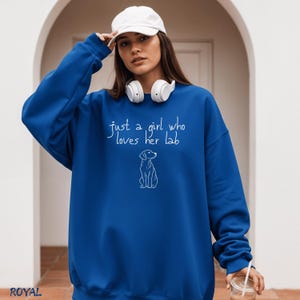 Puede incluir: Sudadera azul real con el texto "just a girl who loves her lab" y un dibujo en blanco de un perro. La sudadera tiene cuello redondo y mangas largas. Tambi&eacute;n se ven una gorra de b&eacute;isbol blanca y unos auriculares.