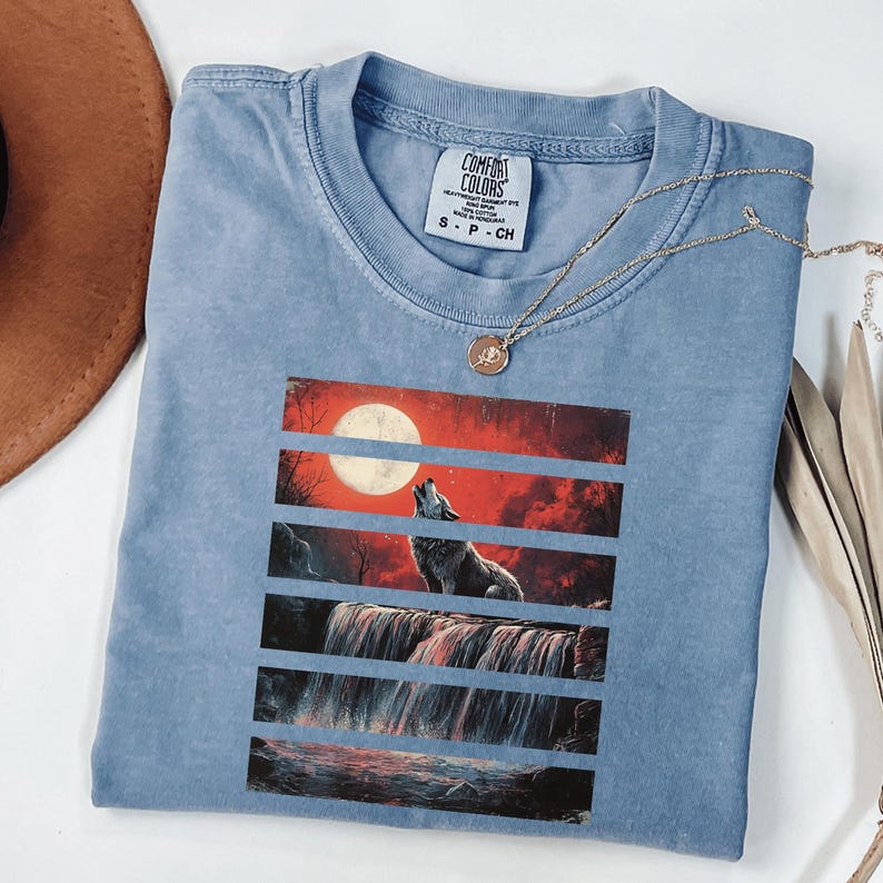 May include: A light blue t-shirt with a graphic print of a wolf howling at the moon in front of a waterfall. The graphic is in a layered style with red, orange, and black stripes. The t-shirt has a tag that says "Comfort Colors" and "S - P - CH".