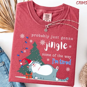 Puede incluir: Camiseta roja carmes&iacute; con un gato de dibujos animados con gorro de Pap&aacute; Noel, &aacute;rbol de Navidad y texto: "probably just gonna jingle some of the way I'm tired". Un collar plateado est&aacute; sobre la camiseta.