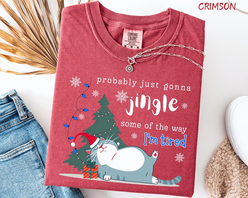 Puede incluir: Camiseta roja carmes&iacute; con un gato de dibujos animados con gorro de Pap&aacute; Noel, &aacute;rbol de Navidad y texto: "probably just gonna jingle some of the way I'm tired". Un collar plateado est&aacute; sobre la camiseta.