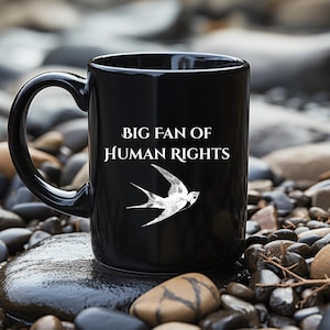 Op de afbeelding: Een zwarte keramische mok met de witte tekst BIG FAN OF HUMAN RIGHTS en een witte grafische illustratie van een vogel in vlucht. De mok heeft een gebogen handvat en een gladde, glanzende afwerking met duidelijke typografie en een vogel.
